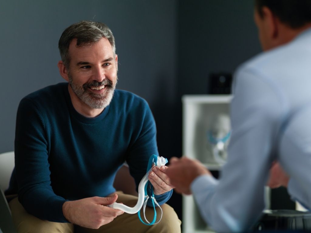 Man in a blue sweater sitting and smiling while holding an F&P Nova Micro mask listening to a clinician