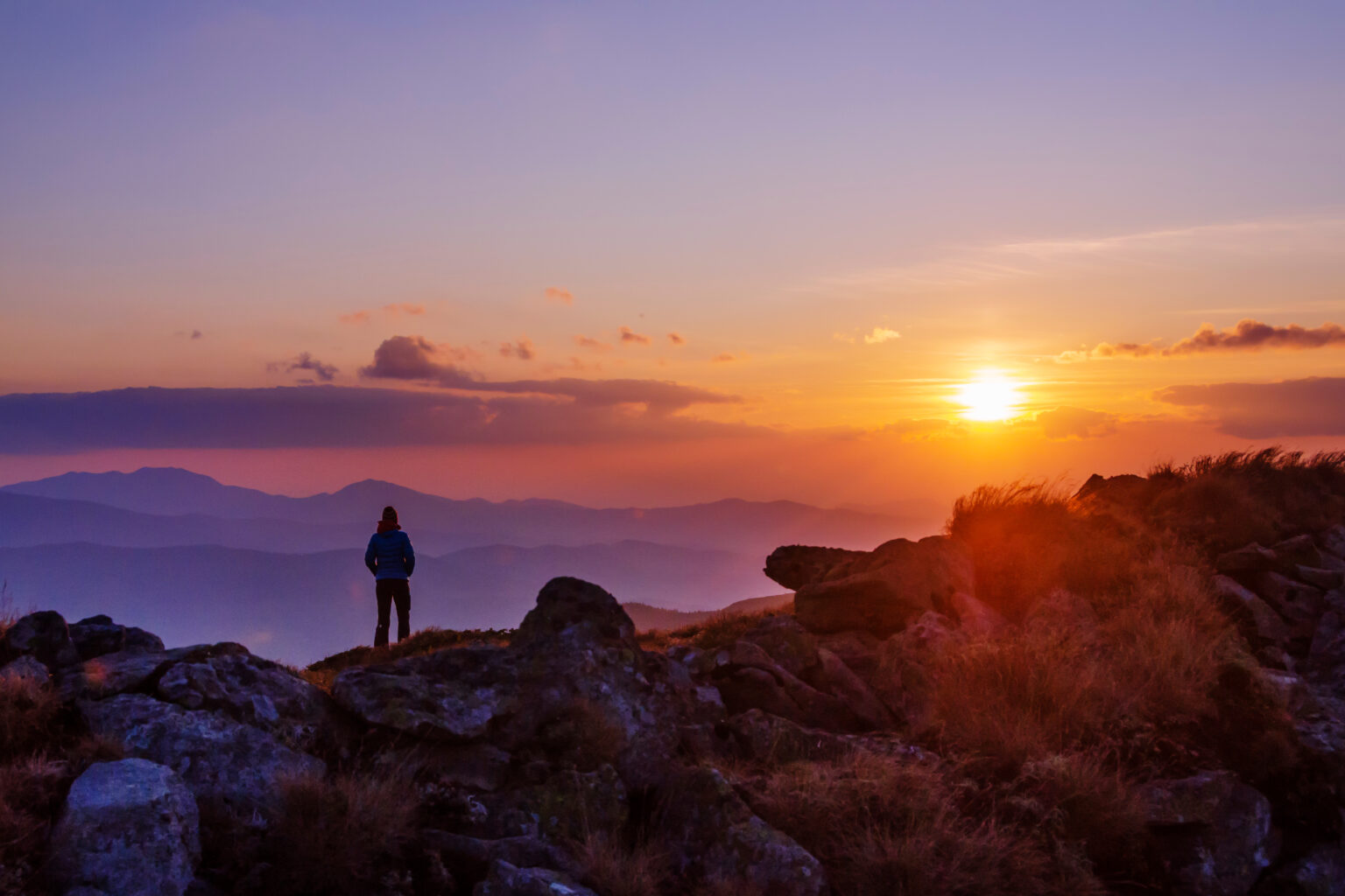 Person standing on a mountain top watching a sunset over a mountain range