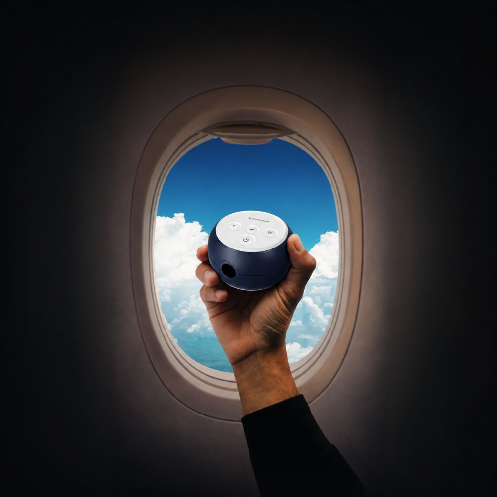 A hand holding a blue Transcend Micro 510 CPAP machine in front of an airplane window, with a view of blue sky and clouds outside.